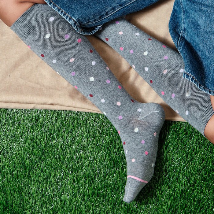 Gray knee-high socks with colorful polka dots and a pink cuff, resting on a fabric surface and grass.
